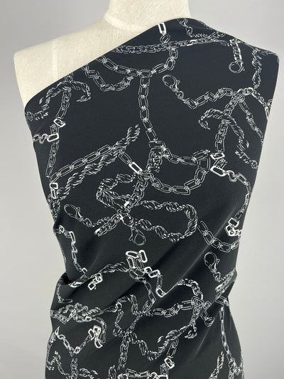 A black fabric draped over a mannequin, featuring a pattern of interconnected silver chains. The intricate design creates a dynamic and textured look against the dark background. This versatile fabric is arranged asymmetrically, highlighting its fluidity and structure. Introducing Deluxe Print - MonoLinks - 150cm by Super Cheap Fabrics.