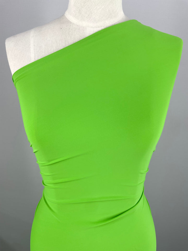 A close-up image of a mannequin wearing a vibrant green, one-shoulder dress made of smooth Nylon Lycra - Green Flash - 160cm fabric. The dress features a single sleeve and is form-fitting, draping elegantly over the mannequin's torso. The background is a plain, light gray color. This stunning fabric is brought to you by Super Cheap Fabrics.