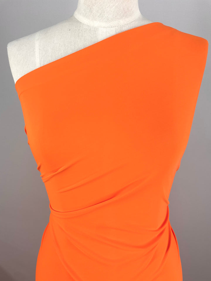 A mannequin is dressed in a vibrant, exotic orange one-shoulder dress made from Super Cheap Fabrics' Nylon Lycra. The material, which measures 150cm wide, is ruched at the sides to create a draped effect. The plain grey background enhances the bright color and stylish design of the dress.