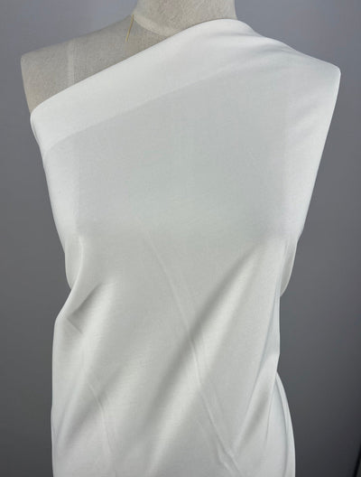 A white, one-shoulder draped woven fabric covers the upper torso of a mannequin, set against a plain gray background. The fabric appears slightly translucent and has a smooth texture. This fabric is the Light Weight Stretch Bengaline - White -150cm by Super Cheap Fabrics.