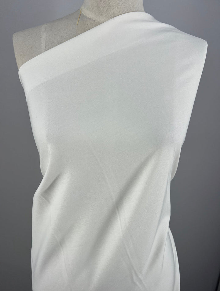 A white, one-shoulder draped woven fabric covers the upper torso of a mannequin, set against a plain gray background. The fabric appears slightly translucent and has a smooth texture. This fabric is the Light Weight Stretch Bengaline - White -150cm by Super Cheap Fabrics.