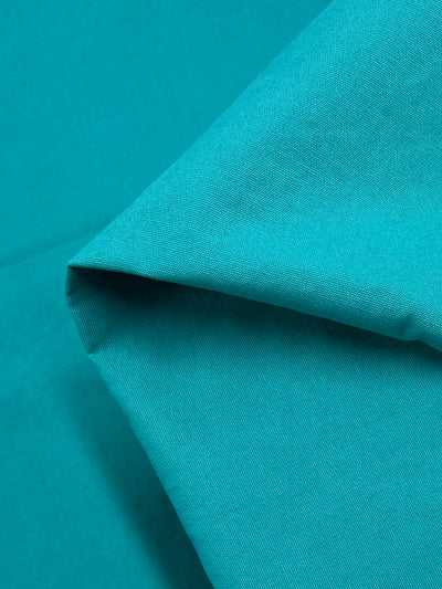 A close-up shot of a folded, bright aqua fabric. The image captures the texture and smooth surface of the 100% cotton material, with visible folds and creases. The lighting highlights the fabric’s weave, emphasizing its vibrant color. This is "Cotton Canvas - Bright Aqua - 147cm" by Super Cheap Fabrics.