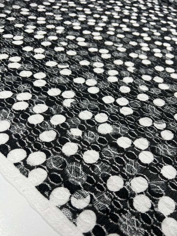 A close-up of Super Cheap Fabrics' Evening Lace - Lemony - Black & White - 142cm. The lightweight fabric features alternating white circular dots on a black background, creating a repetitive and geometric design. The texture of the fabric is clearly visible, adding depth to the image.