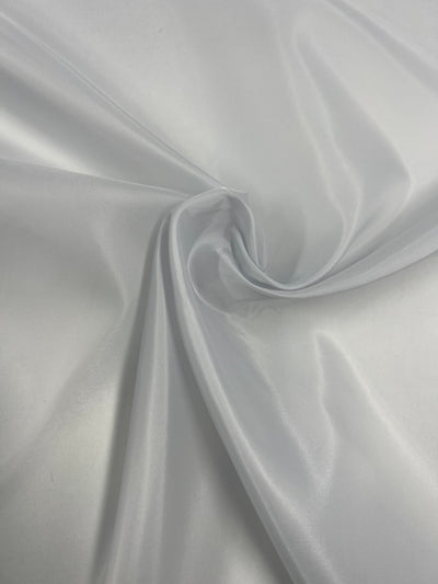 A close-up photograph of shiny, smooth white polyester lining. The fabric is slightly twisted in the center, creating a gentle swirl effect that highlights its glossy texture and delicate folds. This lightweight Lining - Silver - 120cm from Super Cheap Fabrics appears soft and reflective under light.