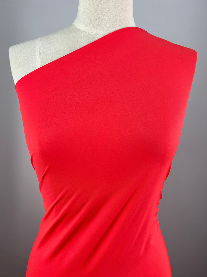 A close-up of the Super Cheap Fabrics' Nylon Lycra dress in Fiery Red, displayed on a mannequin. The four-way stretch fabric appears smooth and form-fitting, with the left shoulder and collarbone exposed. The neutral background emphasizes the vibrant color and sleek design of this 155cm wide dress.