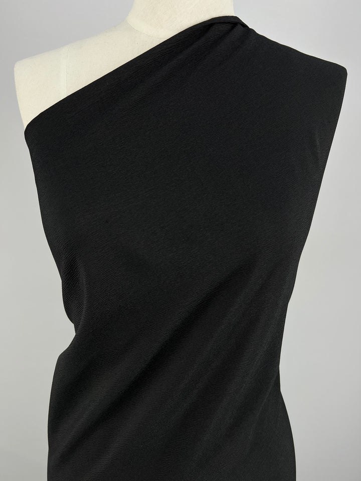 A mannequin adorned with a sleek, one-shoulder outfit stands against a simple gray backdrop. The fabric, made from Super Cheap Fabrics' Ramie Linen in black, appears smooth and fitted, highlighting the garment's elegant design.