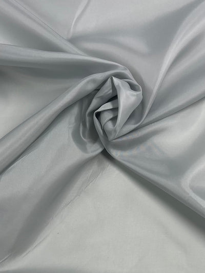 A close-up image of light gray satin fabric. The lightweight fabric is slightly crumpled and folded, creating a swirling pattern in the center. The smooth and glossy texture of the satin is visible, highlighting its metallic sheen—perfect for adding a garment accent like Super Cheap Fabrics' Lining - Northern Droplet - 112cm.