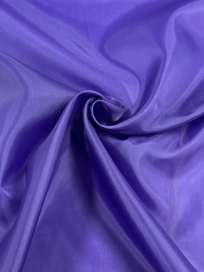 A close-up view of a piece of smooth, glossy Lining - Purple - 112cm from Super Cheap Fabrics. The fabric is laid out with gentle folds and a swirl pattern in the center, showcasing its shiny texture and rich color. The vibrant accent of this satin makes it perfect as a lightweight lining fabric.