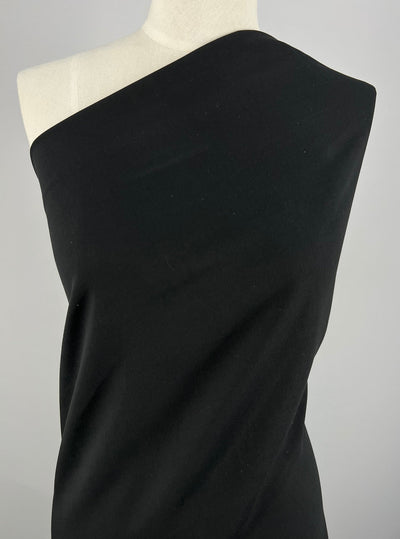 A mannequin adorned with Milano Ponte - Black - 160cm fabric from Super Cheap Fabrics is draped over one shoulder, creating an asymmetrical look. The background is plain and gray, highlighting the elegant draping of the wrinkle-resistant fabric.