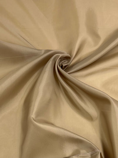 A swirl of beige satin fabric is spread out, showcasing its smooth and shiny texture. This lightweight lining fabric, Lining - Fennel Seed - 112cm by Super Cheap Fabrics, is elegant and has a slight sheen that reflects light, emphasizing its luxurious appearance and making it an ideal garment accent.